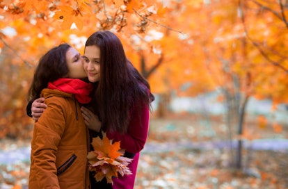 Beautiful teenager daughter kisses mother on the cheek for a walk in the park in fall. A kid in a sc...