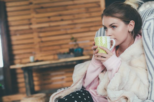 Young beautiful attractive woman in cozy clothes sitting alone in chair. Hold cup with hot drink and...
