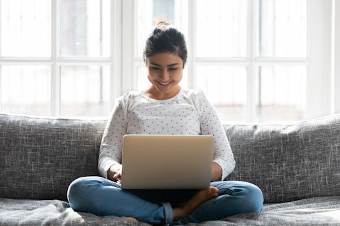 Smiling Indian woman using laptop, sitting on comfortable couch at home close up, happy girl chattin...
