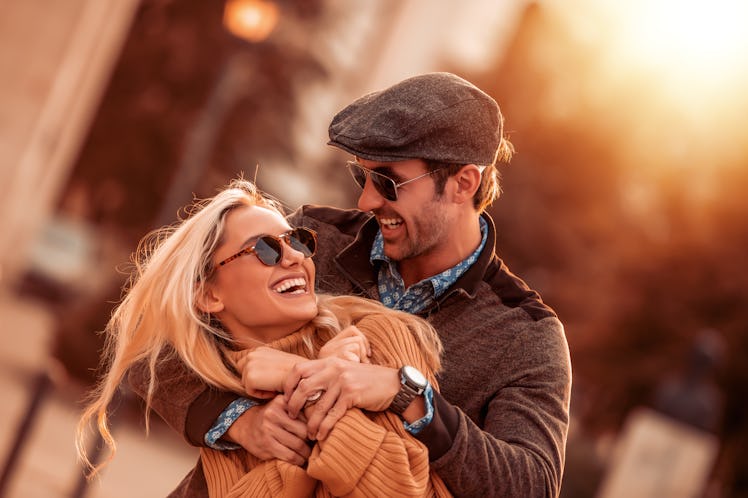 A couple in fall clothes and sunglasses hugs and laughs in the sunshine on Thanksgiving.