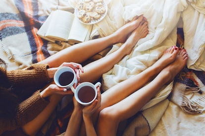 Two friends sit on a bed with cups of tea, blankets, and cookies in the fall.