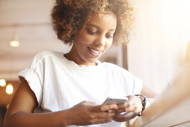 Portrait of good-looking African female student dressed casually holding mobile phone, typing messag...