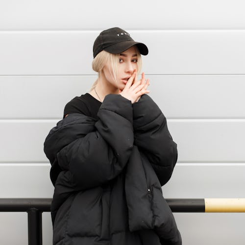 Fashion pretty model girl in fashionable black winter clothes with a cap near a modern metal wall