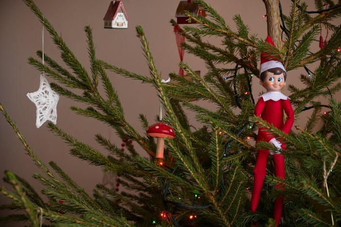 an elf on the shelf perched on a decorated christmas tree with lights.