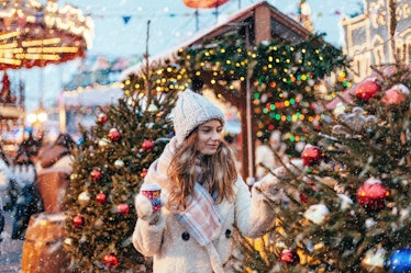 Girl drinking hot coffee while walking in Christmas market decorated with holiday lights in the even...