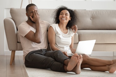 Young overjoyed happy african american family couple sitting on floor mat with computer on knees at ...