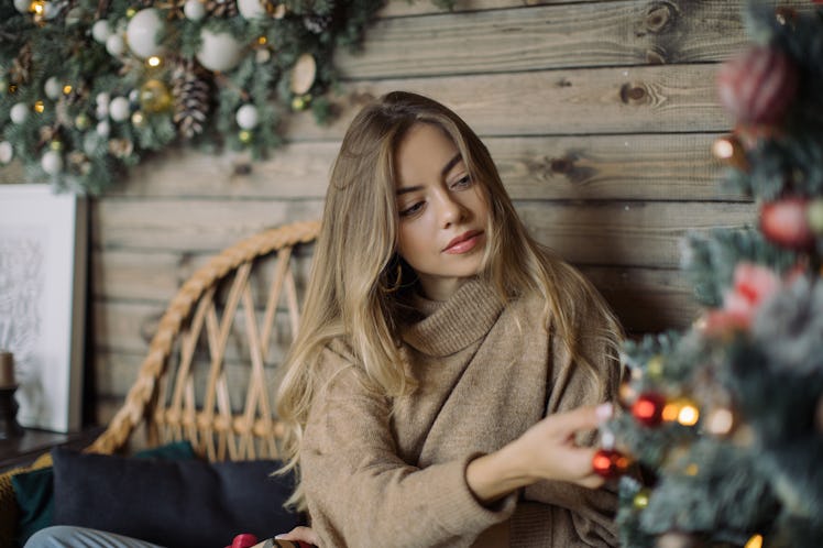 A woman sits next to a Christmas tree in her home while hanging an ornament.