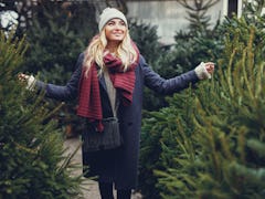 A blonde woman smiles and walks amongst a row of Christmas trees.
