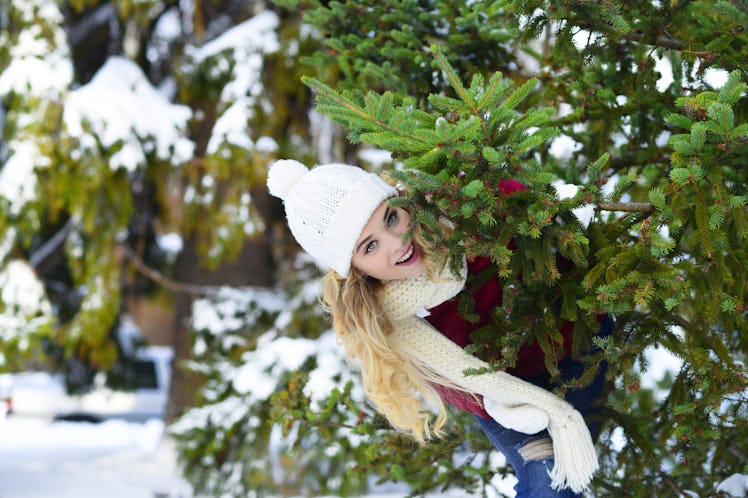 A blonde woman smiles and peaks out from behind a Christmas tree at a farm in the winter. 