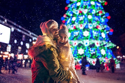 A couple enjoys a fun holiday date outside, hugging in front of a Christmas tree that is all lit up.