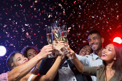 A group of friends laughs and toasts their champagne glasses while confetti falls on New Year's Eve.