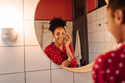 Brown-eyed woman in great mood looks in bathroom mirror and applies cream