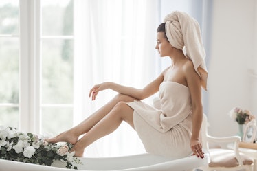 A woman sits by a bathtub with flowers, in a cream-colored bath towel and towel around her hair look...