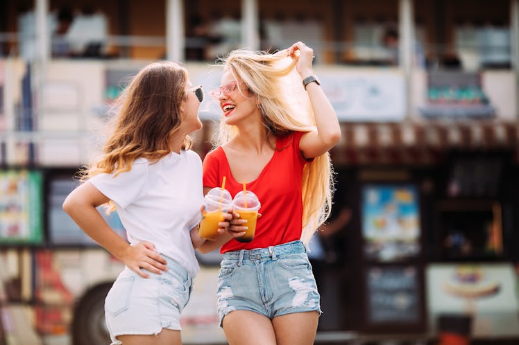 Two stylish girls laugh while they hold drinks in their hands and walk through an amusement park. 