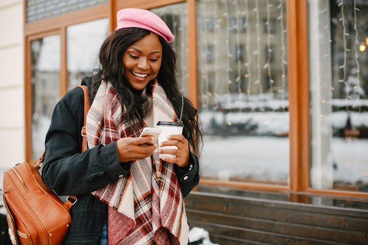 A fashionable woman walks around a city with a backpack, scarf, and pink hat, looking at her phone a...