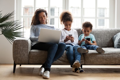 A mom sits on the couch with her two children, all using devices. 