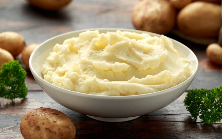 Mashed potatoes in a white bowl are placed on on wooden, rustic table with potatoes surrounding it.