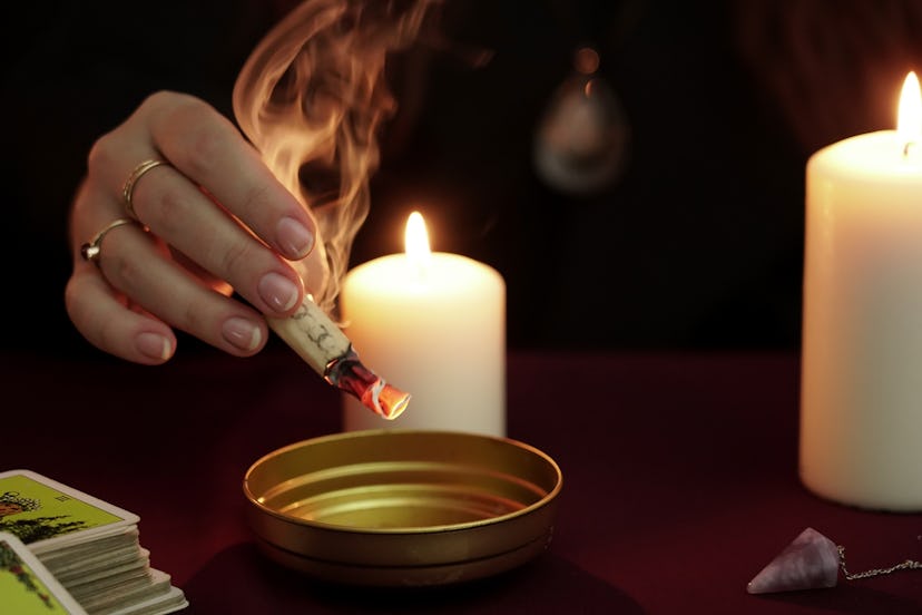 Witch is fortune teller in black mantle holding burning paper with spell over bowl. Tarot cards, ame...