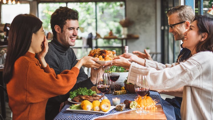 A group of four friends smiling and laughing, passing the turkey to each other, while enjoying Thank...