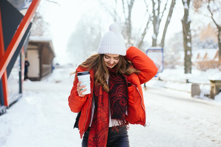 A smiling girl walks through the snow in a red, puffy jacket, with a to-go cup in her hand.