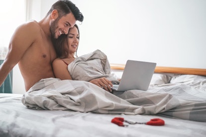 Happy couple watching on computer while lying on bed under blanket - Young lover watching role games...