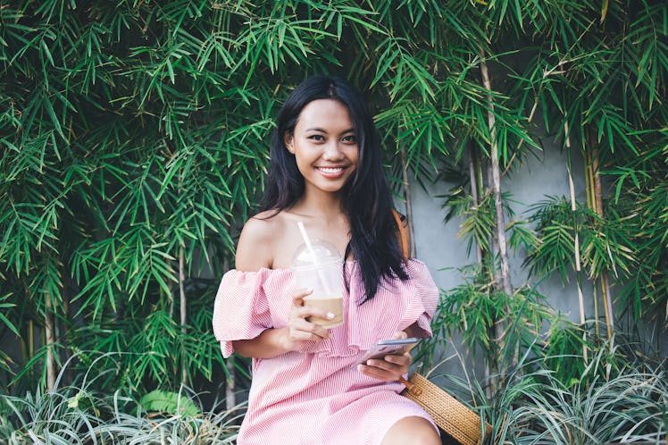 Cheerful pretty young Asian woman in pink off-shoulder dress drinking takeaway coffee and texting on...