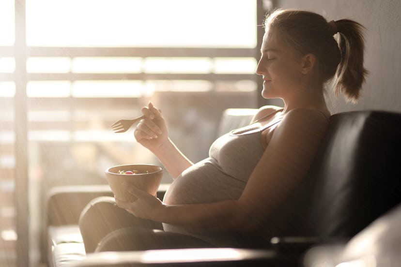 Portrait of pregnant woman sitting on a sofa. She is holding wooden fork and salad bowl. 