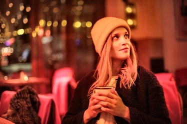 A blonde girl sits in a bar with red lighting in the winter with a holiday drink in her hands.