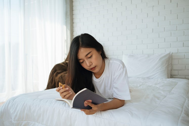 A woman lays on her bed with a gingerbread cookie and book in her hands.