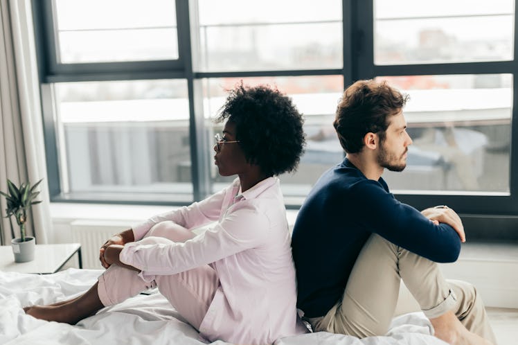 Close-up of an upset interracial couple sitting in bed, back to back, after the quarrel, turning awa...