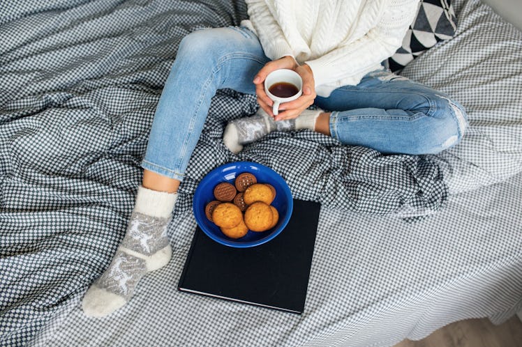 A woman sits on a bed with a blank and white patterned blanket, and eats gingerbread cookies and hol...