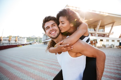 A guy gives his girlfriend a piggyback ride while she kisses his cheek on a sunny day.