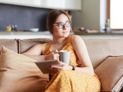 A young woman wearing glasses and a yellow patterned dress uses a tablet while relaxing at home with...