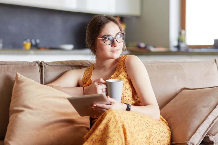 A young woman wearing glasses and a yellow patterned dress uses a tablet while relaxing at home with...