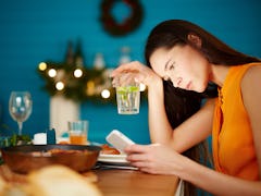 Attractive young female sitting alone at table holding glass and using smartphone during dinner part...