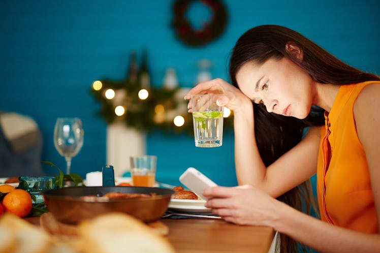 Attractive young female sitting alone at table holding glass and using smartphone during dinner part...