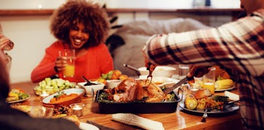 A woman laughs holding a drink in hand while sitting at a Thanksgiving dinner table and the guy acro...