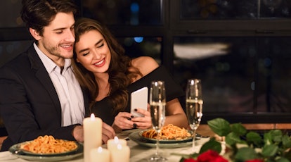A happy brunette couple looks down at photos on a phone during a romantic dinner with champagne and ...