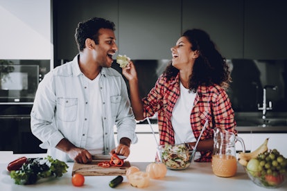A happy couple cooking makes salad at a white kitchen counter, while the girl laughs and feeds her b...