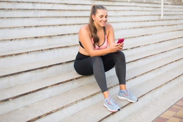 Young cheerful plus size woman in sporty top and leggings sitting on stairs happily using cellphone ...