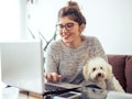 A brunette woman wearing glasses and her hair in a bun smiles and types on her computer with her pup...