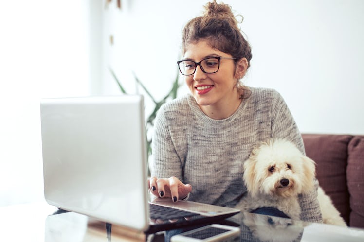 A brunette woman wearing glasses and her hair in a bun smiles and types on her computer with her pup...