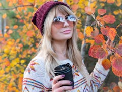 A blonde woman with sunglasses, a colorful sweater, and beanie holds a coffee cup, standing next to ...