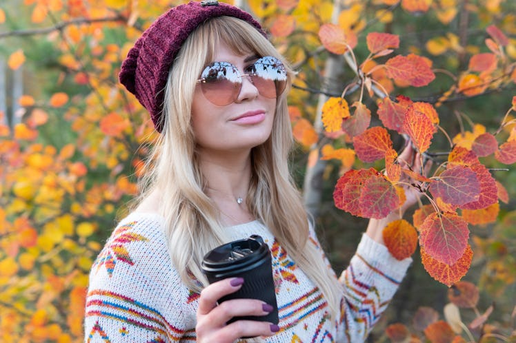 A blonde woman with sunglasses, a colorful sweater, and beanie holds a coffee cup, standing next to ...