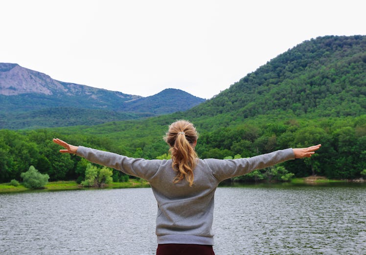 
A bright girl with her arms spread apart is standing against the backdrop of mountains and a lake, ...