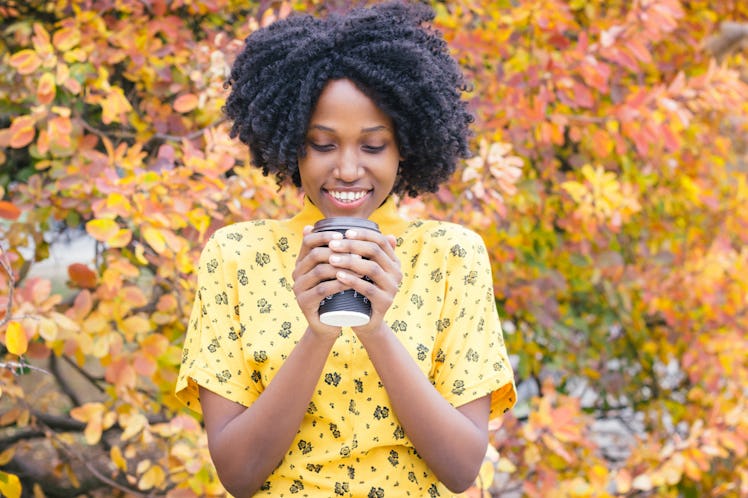 A woman in a yellow T-shirt smiles and looks down at her cup of coffee with yellow foliage in the ba...