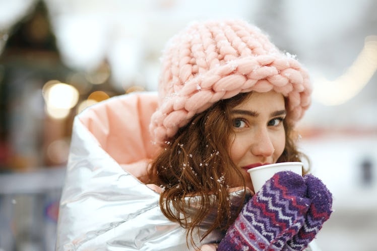 A woman in a metallic coat, pastel pink hat, and purple mittens sips mulled wine at a holiday market...