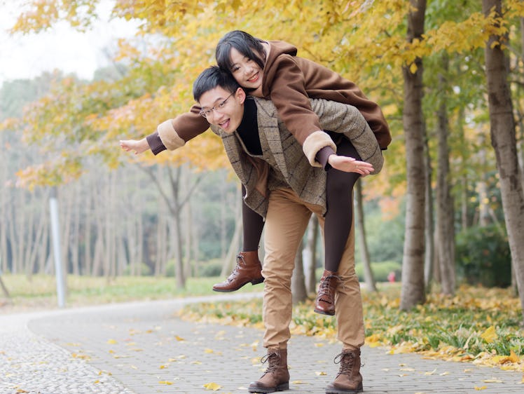 Portrait of smiling young girlfriend piggyback boyfriend during romantic walk in autumn park alley, ...