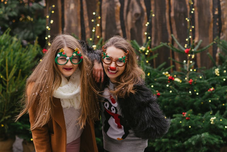 Two girls smile and pose in front of Christmas trees with festive sweaters and glasses on for SantaC...