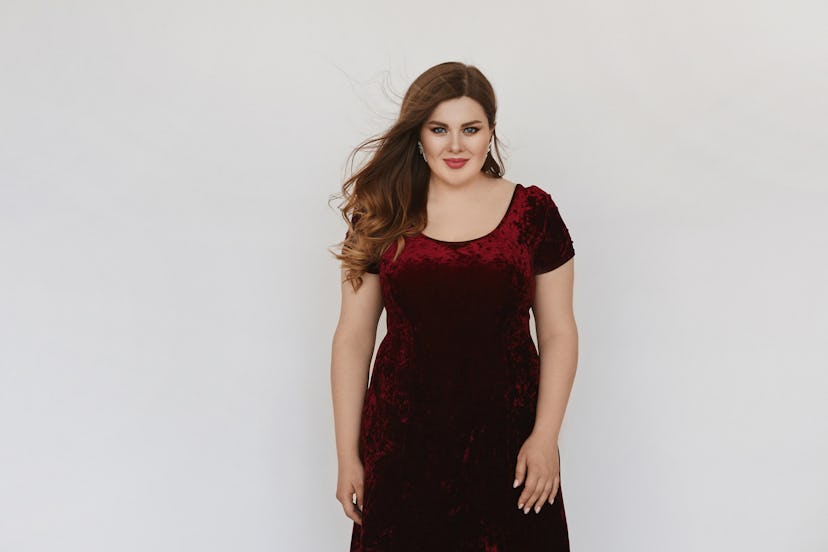 Beautiful plus size model girl in modish red velvet dress isolated at white background. Young fat wo...